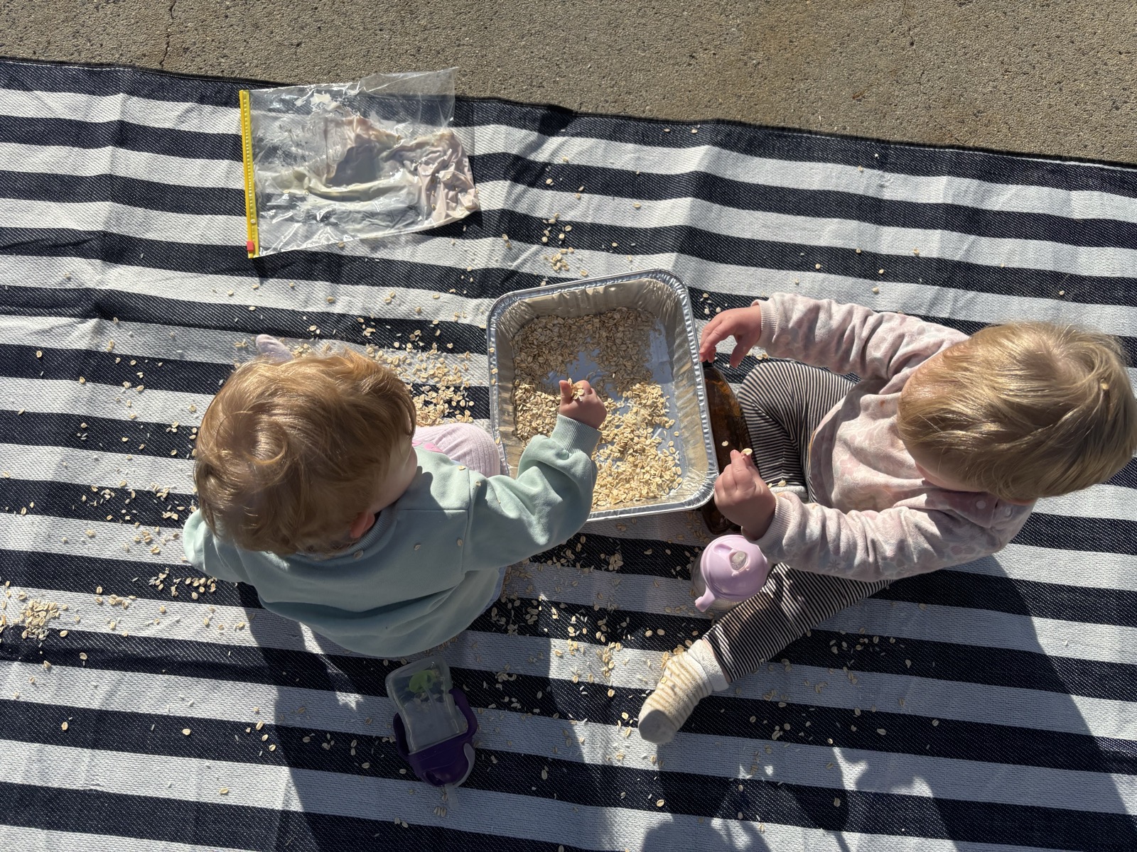 Two children exploring an oats sensory play tray on a picnic rug in the sunshine